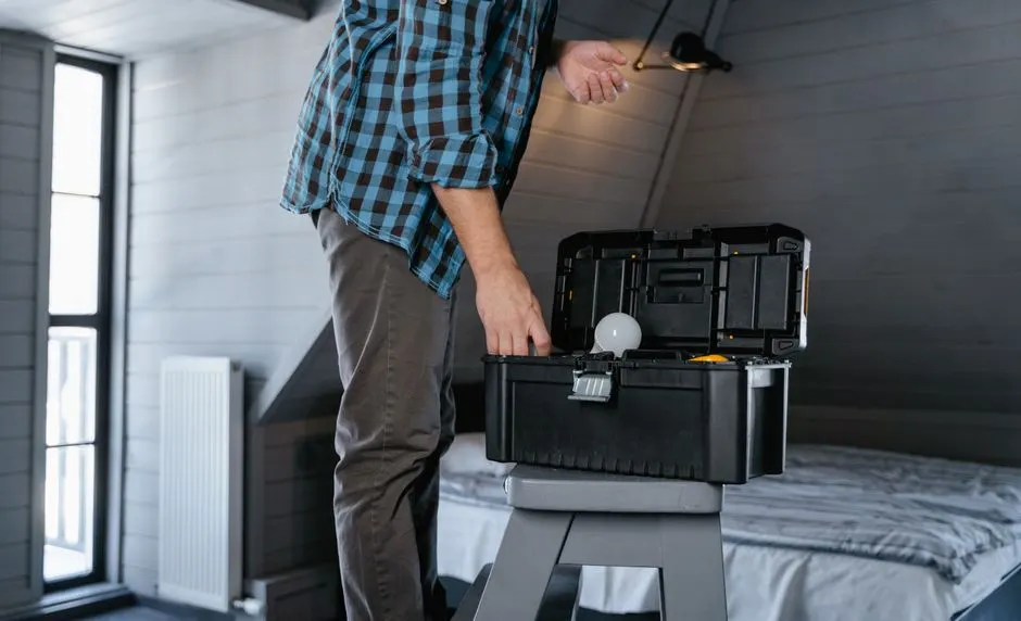 Person in plaid shirt fixing a light bulb using a toolbox in a rustic attic interior.